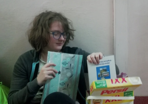 Na podłodze siedzi uczennica w okularach. Obok niej stoi duże pudełko po proszku do prania marki Persil, z którego wyjmuje notatki.